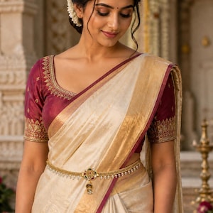 May include: A cream-colored sari with a gold border and a maroon trim. The sari is paired with a maroon blouse with gold embroidery. A gold waist chain with a decorative pendant is worn over the sari. The overall look is elegant and traditional.