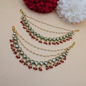 May include: Two gold-toned ear cuffs with a green and white geometric design, accented with small red beads and dangling red clusters. Delicate pearl chains connect the ear cuffs. The background features red and white flowers.