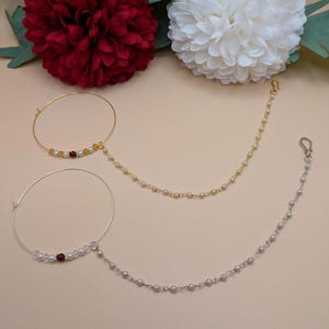 May include: Two gold nose rings with chains. One ring has a red stone and white beads, the other has white beads. Both chains have white beads.