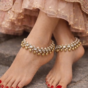 May include: A pair of gold anklets with white and clear stones and small gold bells. The anklets are worn on bare feet with red nail polish. The background is a blurred view of a pink and gold dress.