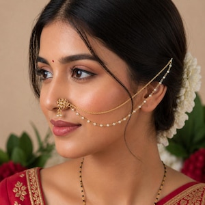 May include: A gold nose ring with a floral design and a dangling pearl. The jewellery is connected to the ear with a delicate chain adorned with small pearls. The woman is wearing a red sari.