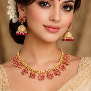Op de afbeelding: Een goudkleurige ketting en bijpassende oorbellen set met rode edelsteen accenten. De ketting heeft een bloemenmotief en de oorbellen zijn klokvormig. De sieraden worden gedragen door een persoon met donker haar en make-up.