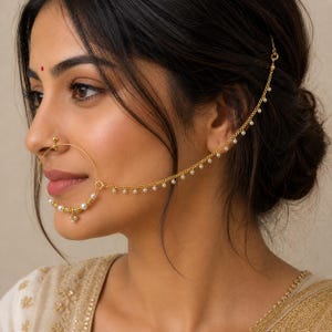 May include: A gold nose ring with a chain that attaches to the ear. The nose ring has a large hoop with small pearls and a dangling charm. The chain is decorated with small pearls. The jewellery is worn on the left side of the face.