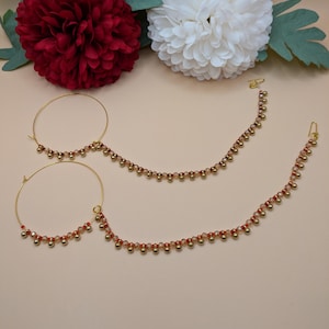 Op de afbeelding: Gouden neusringen met bevestigde kralenkettingen. De kettingen zijn versierd met kleine, ronde, rood-oranje kralen. De neusringen worden gepresenteerd tegen een neutrale achtergrond, met rode en witte bloemenaccenten.