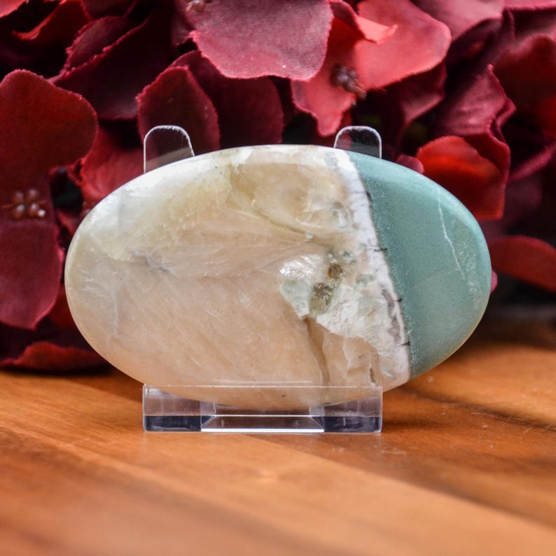 May include: An oval-shaped stone with a polished surface, displayed on a clear acrylic stand. The stone features a blend of colors, including cream, pale green, and white. The background includes red floral elements.