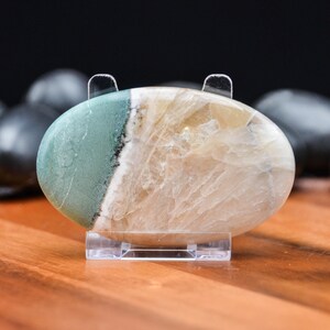 May include: An oval-shaped polished stone, resting on a clear acrylic stand. The stone features a split design with a section of pale green and a section of beige and white. The stone is smooth and has a natural, organic appearance.