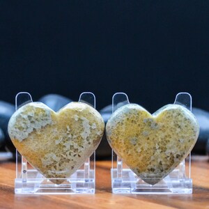May include: Two heart-shaped stones with a yellow and gray pattern, displayed on clear plastic stands. The stones have a textured surface and are set against a dark background, showcasing their unique design.