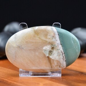 May include: An oval-shaped polished stone with a mix of colors, including cream, green, and hints of brown. The stone is displayed on a clear acrylic stand, showcasing its natural patterns and textures. The stone is approximately 4 inches long.