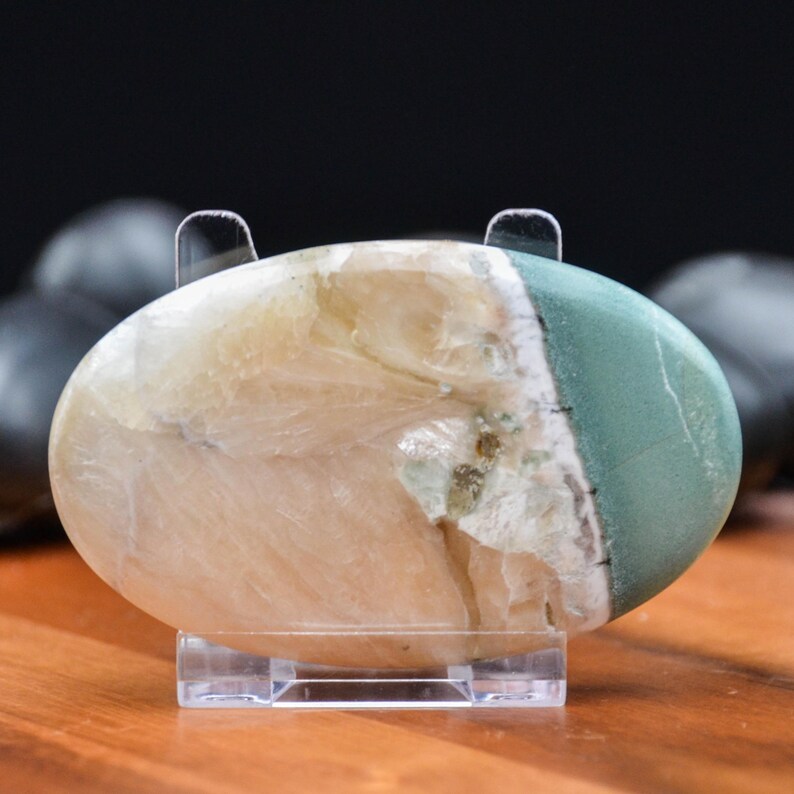 May include: An oval-shaped polished stone with a mix of colors, including cream, green, and hints of brown. The stone is displayed on a clear acrylic stand, showcasing its natural patterns and textures. The stone is approximately 4 inches long.