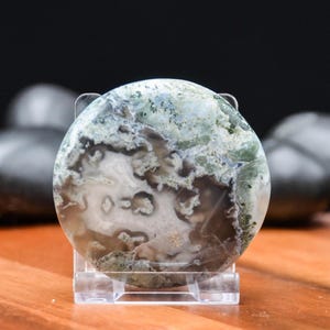 May include: A round, polished gemstone with a unique pattern of white, brown, and green hues. The stone is displayed on a clear acrylic stand, showcasing its natural beauty. The gemstone's surface has a marbled appearance.