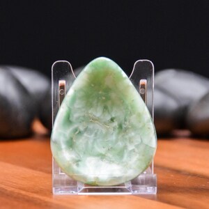 May include: A teardrop-shaped, polished green stone with white marbling, displayed on a clear acrylic stand. The stone has a smooth, glossy surface and varying shades of green. The background is blurred, with dark shapes and a wooden surface.