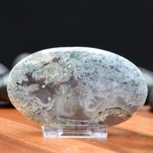 May include: An oval-shaped moss agate stone, displaying a mix of white, green, and gray patterns. The polished stone is mounted on a clear acrylic stand, showcasing its natural textures and colors. The stone's surface has a cloudy appearance.