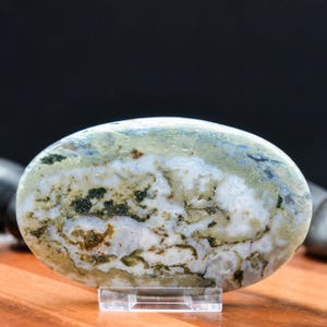 May include: An oval-shaped polished stone with a natural pattern of white, green, and blue hues. The stone is displayed on a clear acrylic stand, showcasing its unique textures and colors. The stone's surface has a marbled appearance.