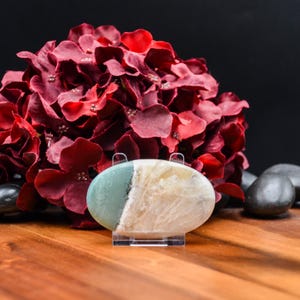 May include: An oval-shaped gemstone, split diagonally with a light green and cream color, is displayed on a clear stand. A large bouquet of deep red flowers is in the background, with black stones and a wooden surface.