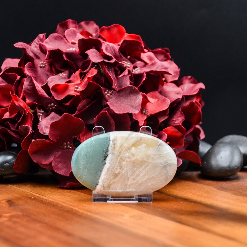 May include: An oval-shaped gemstone, split diagonally with a light green and cream color, is displayed on a clear stand. A large bouquet of deep red flowers is in the background, with black stones and a wooden surface.