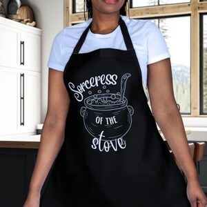 May include: A black apron with a white design of a cauldron with a spoon and the text "Sorceress of the stove".