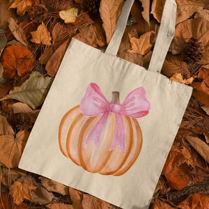 May include: A white canvas tote bag with a pink bow on a pumpkin. The pumpkin is orange and has a stem. The bag is lying on a bed of brown leaves.
