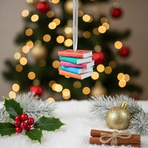 May include: A stack of miniature books, each with a different colored cover, hangs from a white ribbon. The books are suspended in front of a Christmas tree with red and gold ornaments. Holly, berries, and cinnamon sticks are in the foreground.