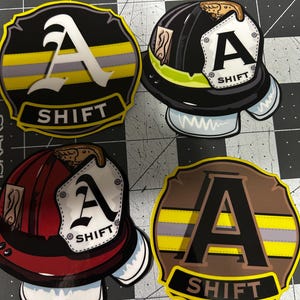May include: Four stickers featuring firefighter-themed designs. Two stickers are shaped like badges with the word "SHIFT" and a large letter "A". The other two stickers are shaped like firefighter helmets, also with the letter "A" and the word "SHIFT".