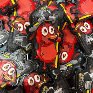May include: A collection of rubberized patches featuring a cartoon dog character with red fur, large eyes with flame-shaped pupils, and a black firefighter helmet. The character holds a yellow pole and wears a black hat with grey accents.