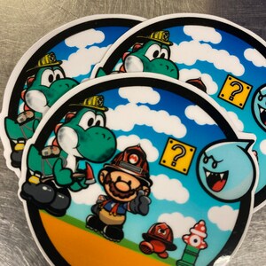 Op de afbeelding: Een sticker met een groen Yoshi-personage dat een bijl vasthoudt, een cartoon brandweerman, een vraagtekenblok en een Boo-spook. De sticker is geplaatst tegen een blauwe lucht met witte wolken.