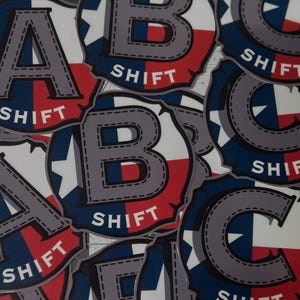 May include: A collection of stickers featuring the letters A, B, and C in a gray, stitched font. Each letter is set against a background of the Texas flag design, with the word "SHIFT" in white.