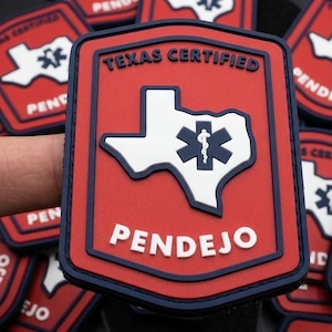 May include: A rectangular red and navy blue morale patch with a white outline of the state of Texas. Inside the state shape is a dark blue Star of Life symbol. The text TEXAS CERTIFIED is at the top and the word PENDEJO is at the bottom in white.