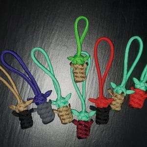 May include: A collection of handmade paracord keychains featuring a green alien-like character. Each keychain has a loop and is crafted in various colors, including green, red, brown, black, purple, and tan. The keychains are arranged on a dark surface.