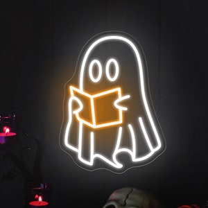 Può includere: Un cartello al neon bianco di un fantasma che legge un libro. Il fantasma ha due occhi e una bocca. Il libro è arancione. Il cartello è su uno sfondo nero con un ramo di albero nero con tre candele accese e un teschio e zucche in primo piano.
