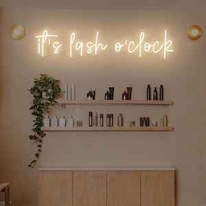 Pode incluir: Interior de um salão de beleza com uma placa de néon que diz "It's lash o'clock". Prateleiras exibem produtos de beleza. O quarto tem uma paleta de cores neutras com armários de madeira e iluminação decorativa.