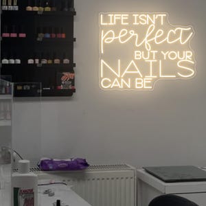 May include: A nail salon interior with shelves of nail polish bottles, a white table with manicure tools, and a neon sign that reads "Life isn't perfect but your nails can be."