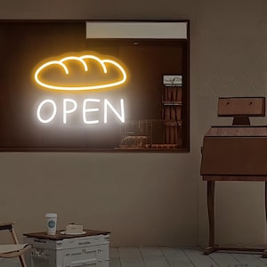 May include: A neon sign with a bread loaf design above the word "OPEN" in white. The sign is in a dark window, with a wooden stand and a small table with a coffee cup in front of it. The sign is likely for a bakery or cafe.