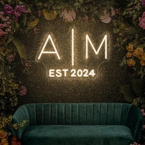 Könnte beinhalten: Ein Neon-Schild mit den Buchstaben "AIM" und dem Text "EST 2024" in weißer Neon-Schrift auf einem dunkelgrünen Hintergrund. Das Schild ist von üppigem Grün und bunten Blumen umgeben.