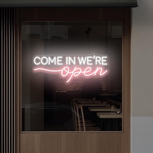 May include: A neon sign in a storefront window reads "COME IN WE'RE open" in white and pink script. The sign is mounted on a dark awning above a glass window, with a glimpse of tables and chairs inside. The sign is from ArtSignDesign.