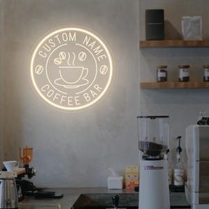 Op de afbeelding: Een oplichtend neonbord voor een koffiebar, met de tekst "CUSTOM NAME COFFEE BAR" rond een koffiekopje en bonenafbeelding. Het bord is bevestigd aan een lichtgrijze muur.