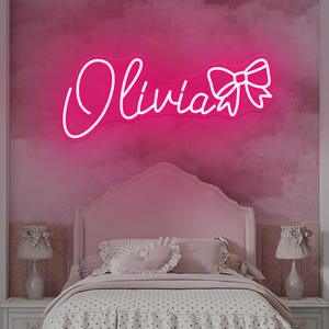 May include: A pink neon sign displaying the name "Olivia" in a cursive font, with a bow accent. The sign is mounted on a pink wall above a bed with pink bedding and a white nightstand. The room has a soft, feminine aesthetic.