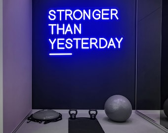 Op de afbeelding: Een sportschoolinterieur met een blauw neonbord met de tekst "STRONGER THAN YESTERDAY". Er is een zwarte yogamat, kettlebells, een foamroller en een grijze oefenbal. De muren zijn donkerblauw en paars.