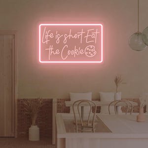 May include: A pink neon sign with the text "Life is short Eat the Cookie" in a rectangular frame. The sign is mounted on a light-colored wall in a cafe setting. The cafe has tables, chairs, and decorative lighting.