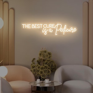Könnte beinhalten: Ein Neonschild mit dem Text "THE BEST CURE is a Pedicure" in einem Spa-Ambiente. Das Schild leuchtet in warmweiß. Der Raum verfügt über zwei geschwungene Sessel, einen kleinen runden Tisch und dekorative Pflanzen.