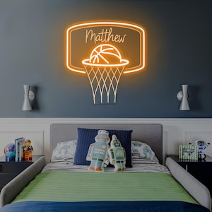 May include: An orange neon basketball hoop sign with the name "Matthew" above the basketball. The sign is mounted on a dark blue wall above a bed with a green and blue comforter. Two robot pillows are on the bed.