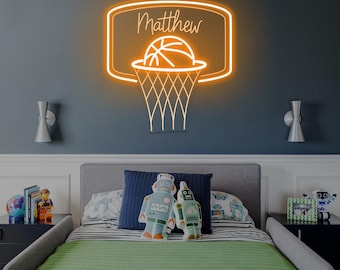 May include: An orange neon basketball hoop sign with the name "Matthew" above the basketball. The sign is mounted on a dark blue wall above a bed with a green and blue comforter. Two robot pillows are on the bed.