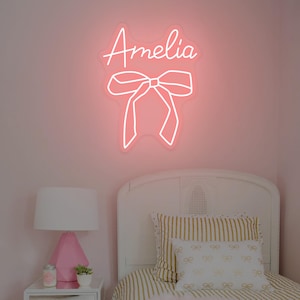 Op de afbeelding: Een roze neonbord met de naam "Amelia" in cursief schrift boven een strik-ontwerp. Het bord is bevestigd aan een roze muur boven een wit bed met gestreepte beddengoed en decoratieve kussens. Een roze lamp en een nachtkastje zijn zichtbaar.
