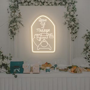 Op de afbeelding: Een oplichtend neonbord in de vorm van een vintage telefoon met de tekst "Leave Us A Message". Het bord staat tegen een witte achtergrond, versierd met bloemstukken, boven een tafelsetting.