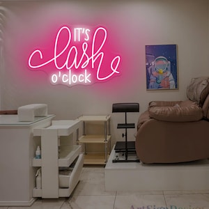 Op de afbeelding: Een roze neonbord met de tekst "It's lash o'clock" hangt aan een muur. De afbeelding toont een schoonheidssalon interieur, inclusief een massagestoel, een nageltafel en een klein kunstwerk van een astronaut.