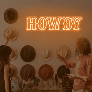 May include: An illuminated orange neon sign that reads "HOWDY" is mounted on a wall. The sign is the focal point, with a collection of cowboy hats in various colors and styles arranged around it. Two people are in the image.