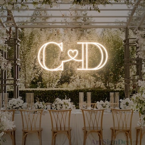 Op de afbeelding: Een bruiloftreceptie met een neonbord met de letters "C&D" en een hartsymbool. De tafel is gedekt met witte tafelkleden, houten stoelen en bloemstukken, wat een elegante sfeer creëert.
