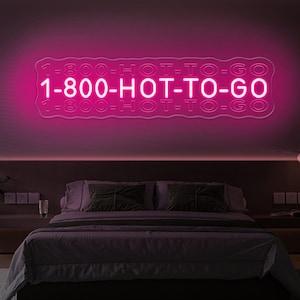 Op de afbeelding: Een roze neonbord met de tekst "1-800-HOT-TO-GO" in een golvend lettertype. Het bord hangt aan een muur boven een bed met een donkerpaarse hoofdeinde.