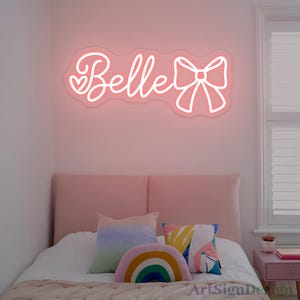 May include: A pink neon sign that reads "Belle" in cursive with a bow and two hearts. The sign is mounted on a pink wall above a bed with decorative pillows. The bed has a pink headboard and a white comforter.