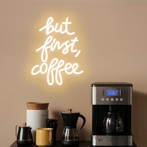 May include: White neon sign with the text "but first, coffee" on a beige wall. A coffee maker, a carafe, and a few coffee pots are on a counter below the sign.