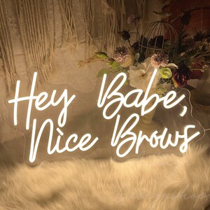 May include: White neon sign with the text "Hey Babe, Nice Brows" on a white fluffy surface.